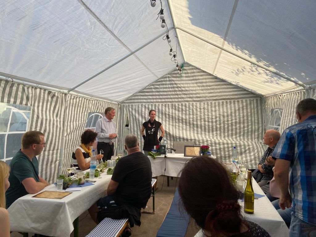 Der Blick auf die Lesebühne: einige Biergarnituren unter einem gestreiften Festzelt, mehrere Leute sitzen und hören der Verlegerin zu. Die steht am Kopfende des Raumes neben dem Winzer.