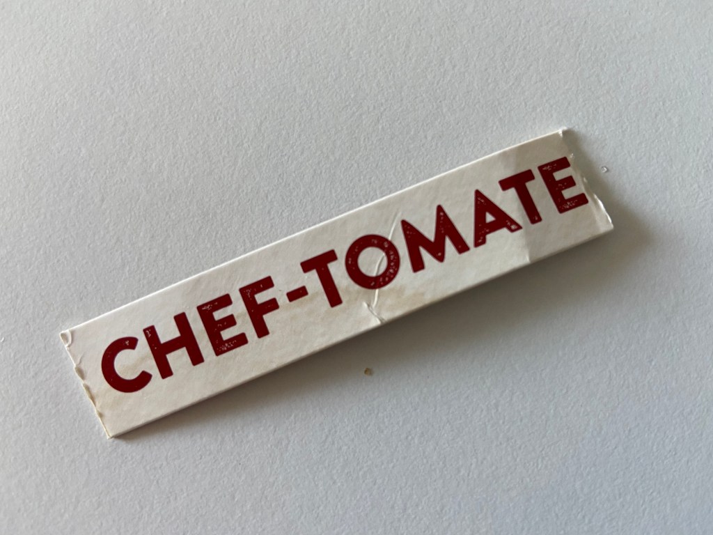 Weißer Hintergrund, darauf ein Pappschnipsel mit der Aufschrift CHEF-TOMATE.