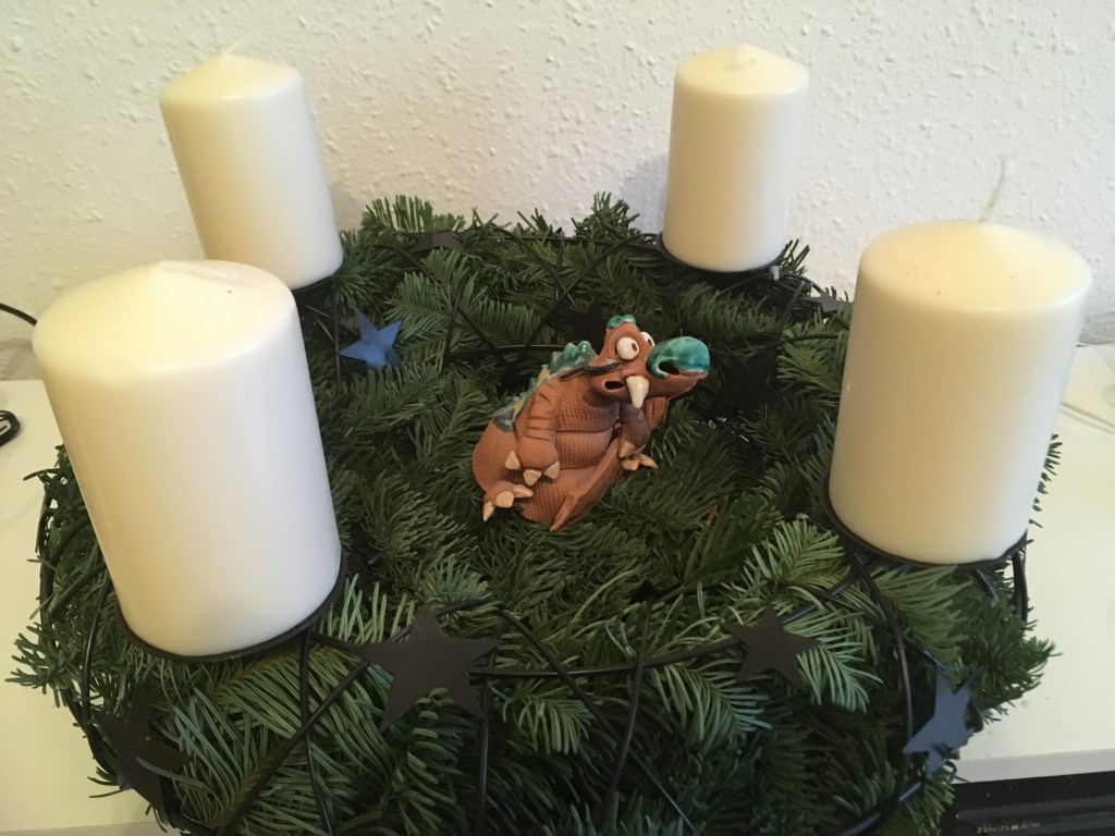 Adventskrank aus Tannengrün in einem Metallkranz mit vier weißen Kerzen. In der Mitte sitzt ein Drache aus Ton.