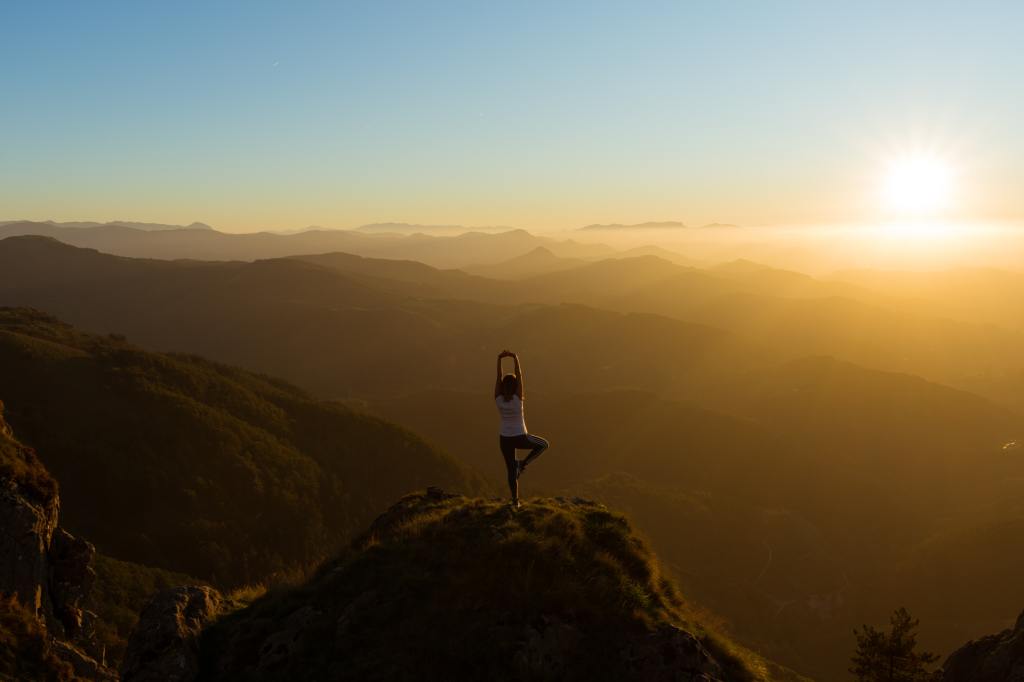 Berggipfel bis zum Horizont, Sonnenaufgang, im Vordergrund steht eine Person mit dem Rücken zum Fotograf in der Yoga-Pose "Baum" auf einem Gipel.