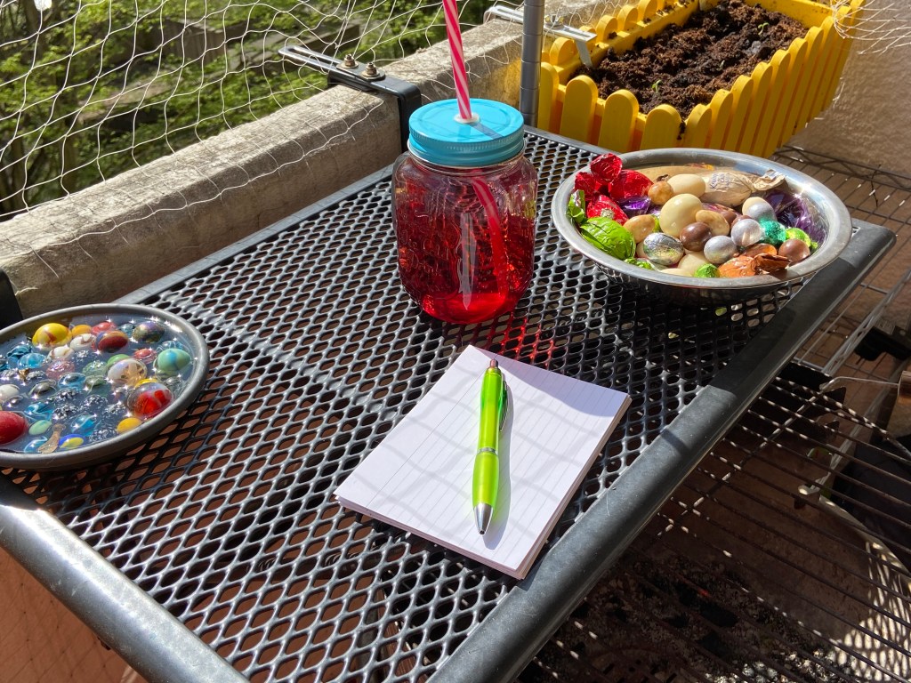 Auf einem Gitter-Balkontisch steht links eine Bienentränke (Wasser und Murmeln in einem Untersetzer), daneben ein Stapel Karteikarten mit einem gelben Kugelschreiber, kalter Früchtetee in einem Glas mit Deckel und Strohhalm, daneben eine Schale mit Ostersüßigkeiten. Im Hintergrund ein Balkonkasten mit Erde.
