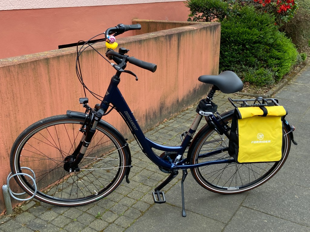 Vor einem Mehrfamilienhaus steht ein dunkelblaues City-Damenrad mit tiefem Einstieg. Auf dem Lenker erkennt man eine gelbe Quietscheente mit Helm. Am Gepäckträger hängt eine gelbe Tasche.