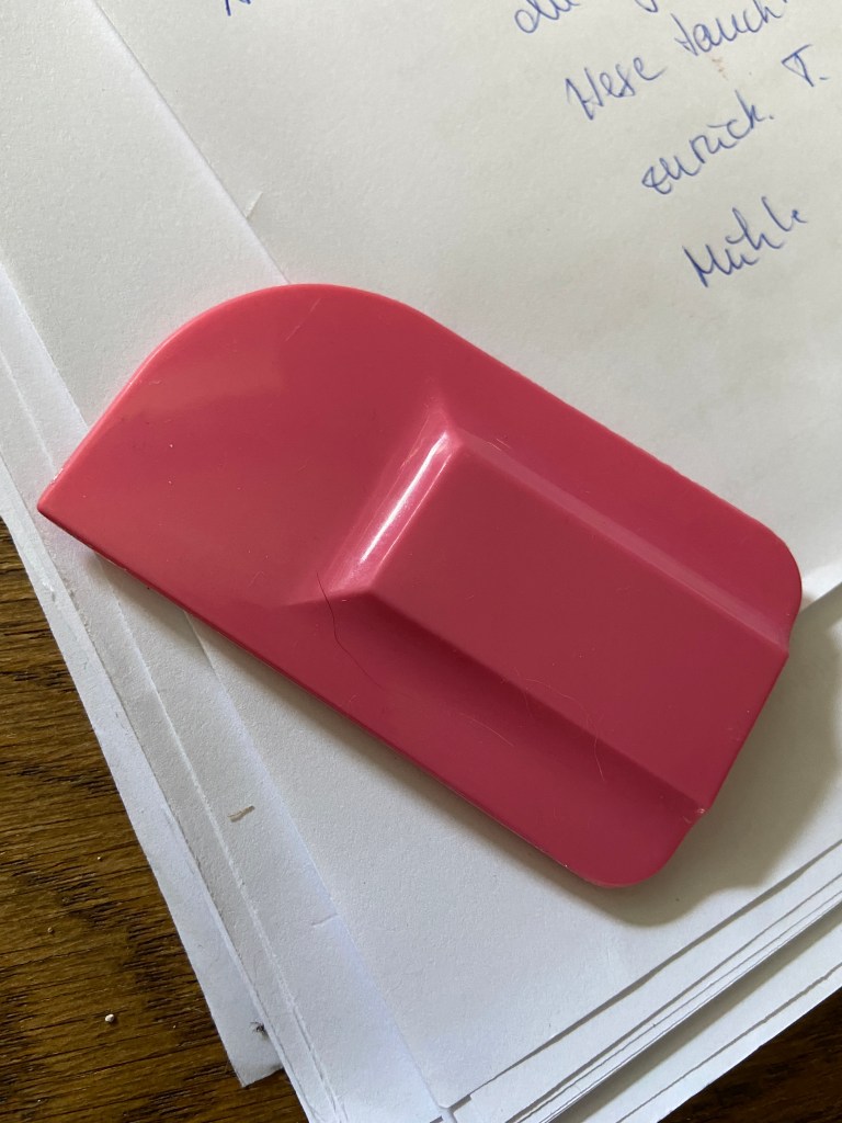 Kopf eines Teigschabers aus rosa Silikon auf einigen vollgekritzelten Blättern Papier.