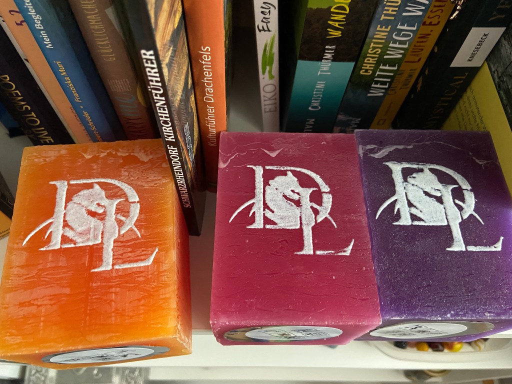 Drei Kerzen (orangefarben, pink, lila) mit Autorinnenlogo aus verschlungenem D und L mit Drachen im D, liegend, im Bücherregal. Man erkennt einige Buchtitel wie "Schwarzrheindorf Kirchenführer" oder "Kulturführer Drachenfels".