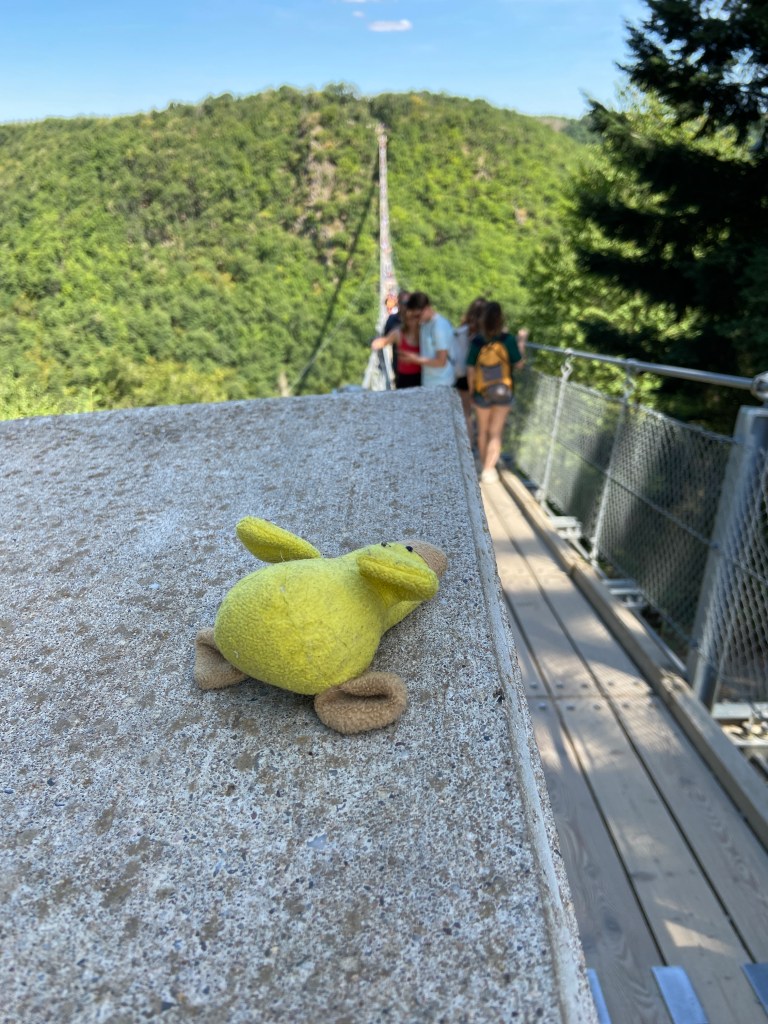 Auf einem Block aus Beton liegt eine kleine gelbe abgenutzte Stoffente, die Keilform hat. Im Hintergrund erkennt man unscharf mehrere Leute, die über eine Hängebrücke gehen, außerdem einen bewaldeten Hang.