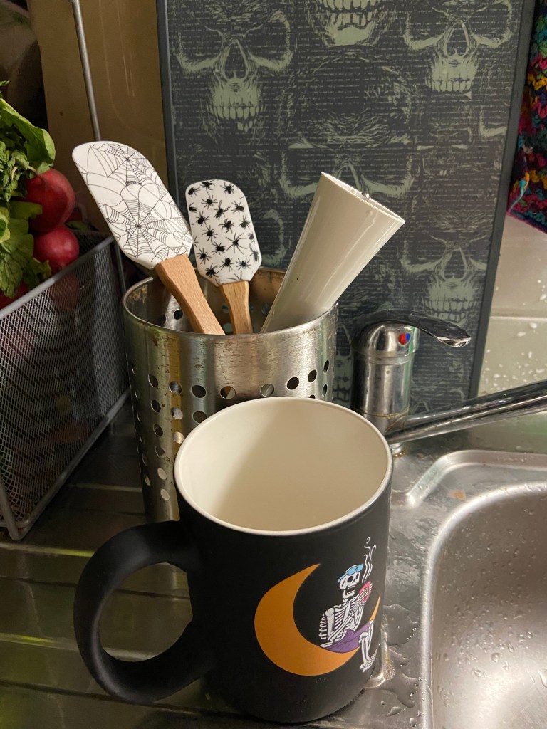 Auf einer Abtropffläche aus Edelstahl steht eine schwarze Tasse mit einem Arbeiterskelett, das auf einer Mondsichel Pause macht. Dahinter, in einem Köcher aus Metall, tropfen zwei Spatel mit Spinnen-Aufdruck ab. An der Wand lehnt ein Schneidebrett, auf dem man verschiedene Totenköpfe in hell auf Grau und Blau sieht. Am Bildrand Radieschen.  