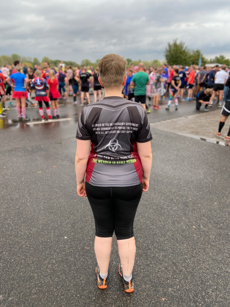 Rückansicht der Autorin daselbst. Sie trägt schwarze Lauftights und ein T-Shirt mit der Rückenaufschrift: "By the order of the UK Emergency Government, you are commanded to provide the wearer with all necessary military supplies. IF YOU CAN READ THIS TEXT, THE WEARER IS STILL HUMAN."
Im Hintergrund viele Läufer im Startbereich, allgemeine Aufregung, alles ist schon ziemlich nass.