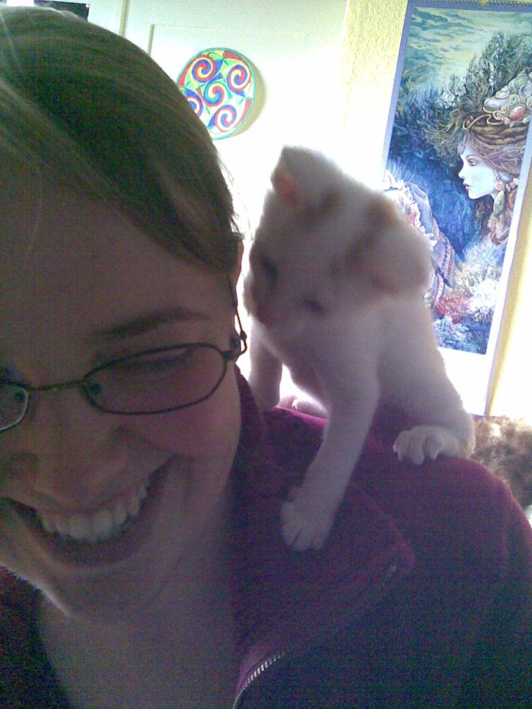 Halb im Bild ist eine junge Frau mit Brille und blonden langen Haaren, lachend. Sie trägt einen magentafarbenen Sweater. Auf ihrer Schulter sitzt ein winziger, beinahe komplett weißer Kater mit ein paar roten Zeichnungen an Kopf und Ohren. Das Bild ist grobkörnig und unscharf.