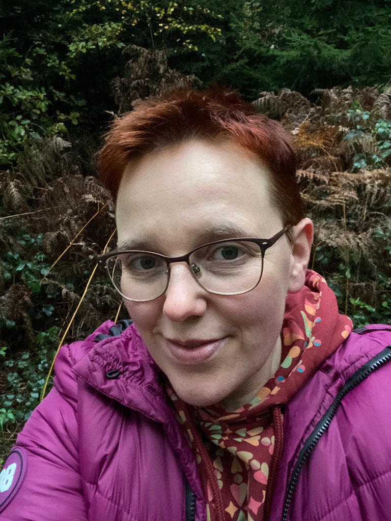 Selfie im Wald. Im Hintergrund dunkles, herbstliches Grün. Im Vordergrund eine sichtlich müde Autorin: Rote Pumuckl-Frisur, blasse Haut mit Falten und Sommersprossen, große Brille mit Drahtgestellt, ungepflegte Augenbrauen, Lächeln. Die Autorin trögt eine violette Allwetterjacke und einen weinroten Sweater mit buntem geometrischem Aufdruck, von dem man nur die Kapuze aus dem Jackenkragen herausragen sieht.