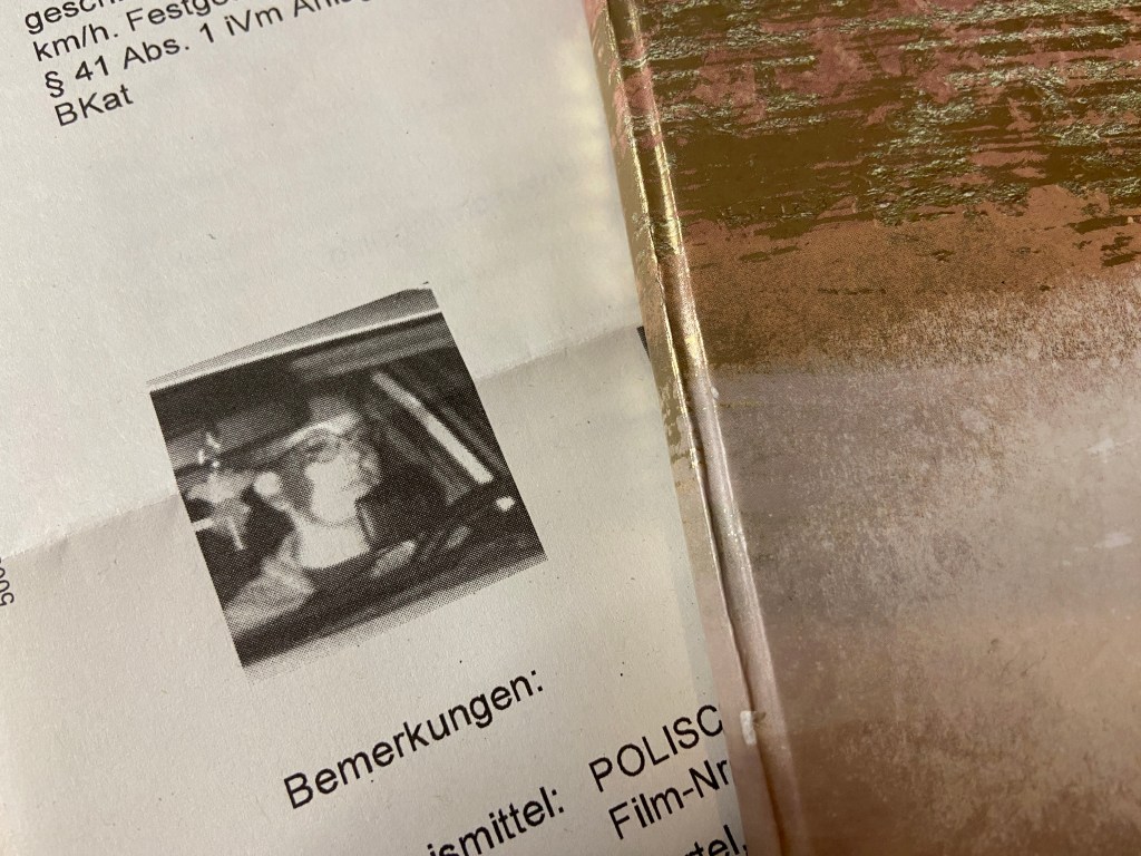 Ausschnitt aus einem amtlichen Schreiben, im Wesentlichen sieht man den Schwarzweiß-Abdruck eines Fotos. Darauf zu sehen ist die Autorin am Steuer eines Autos, konzentriert fahrend. Ebenfalls im Bild sind ein Navi und ein Stofftier, das vom Rückspiegel baumelt.