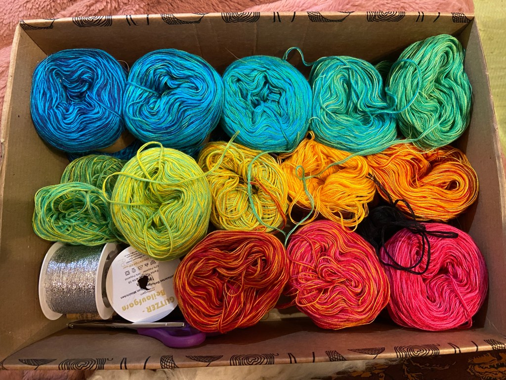 Karton mit mehreren kleinen Garnknäueln in unterschiedlichen Stadien des Verbrauchs. Von oben links (blau) bis unten rechts (rot-pink) insgesamt 13 Knäuel. Unten links in der Box außerdem zwei Spulen mit Beilaufgarn, eines davon silberfarben, eine Bastelschere und eine schlichte Häkelnadel. Schwarzer Faden ragt zwischen den anderen Knäueln hervor.