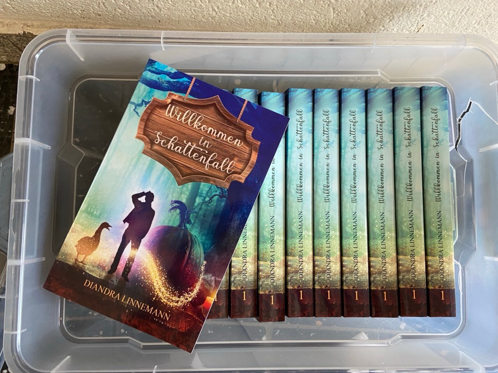 In einer Plastikkiste sieht man ein knappes Dutzend Bücher mit dem Schriftzug "Diandra Linnemann: Willkommen in Schattenfall" auf dem Rücken, ein Exemplar liegt links auf der Kiste. Das Cover zeigt neben Titel und Autorinnennamen die Silhouetten von einer Gans, einem ratlosen jungen Mann, und einem Kürbis. Um den Kürbis winden sich magische Funken, im Hintergrund sieht man einen Herbstwald.