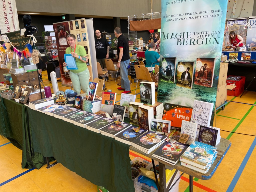Messestand - langer schmaler Biertisch, größtenteils mit einer dunkelgrünen Decke abgedeckt. Darauf, dicht gedrängt, diverse Bücher und etwas Deko und Werbematerial. Im Hintergrund ein Roll-Up: "Magie hinter den sieben Bergen". Links im Bild sieht man meine Standnachbarin Anja Bagus in Aktion.