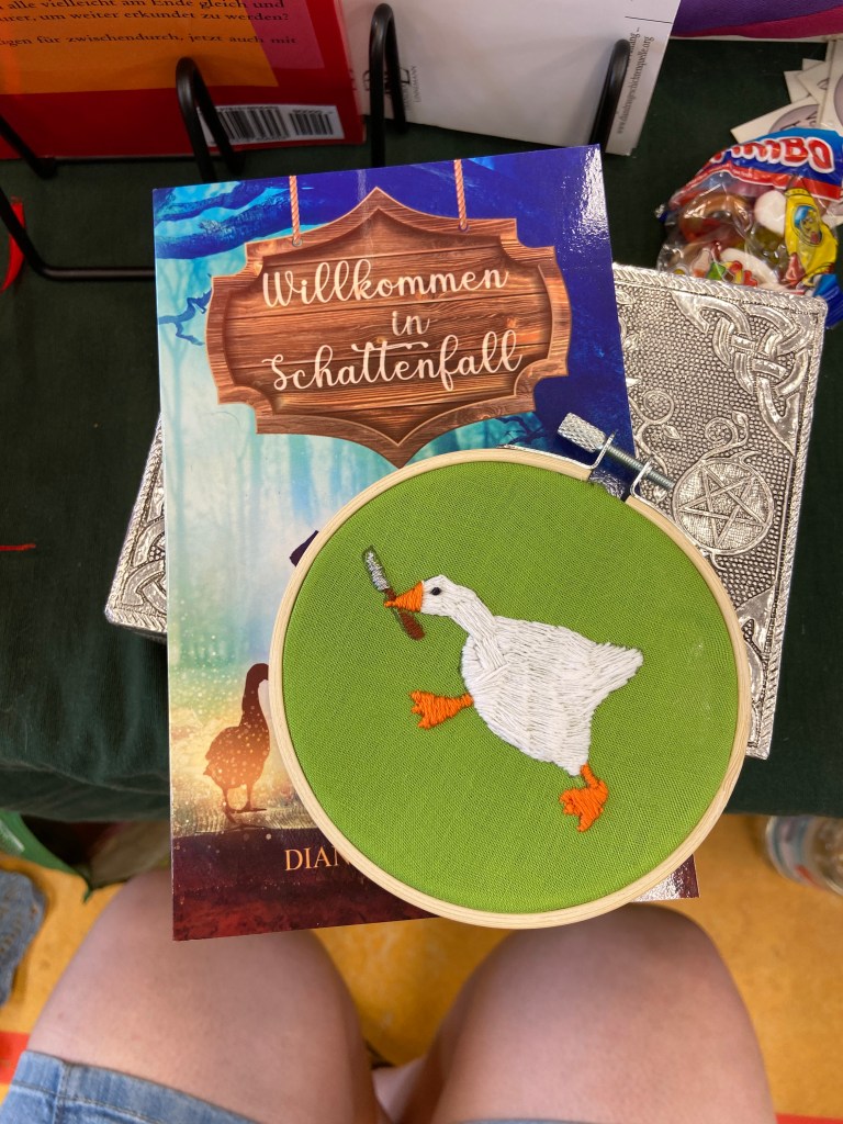 Auf dem Buch "Willkommen in Schattenfall" (Herbstfarben und Hellblau, Schriftzug auf Holzplakette, Silhouette einer Gans links zu sehen) liegt ein Stickrahmen. In diesen Rahmen ist apfelgrüner Stoff gespannt, darauf eine gestickte Gans in Angriffshaltung mit einem Messer im Schnabel. Außerdem sieht man Autorinnenknie.