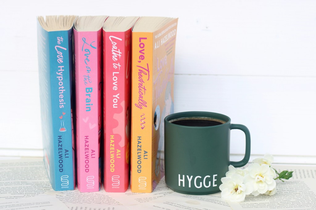 Auf hellem Untergrund sieht man vier fröhlich-bunte Taschenbücher nebeneinander stehen, die Rücken zeigen zum Betrachter. Rechts daneben steht ein tannengrüner Becher mit schwarzem Kaffee, auf dem HYGGE steht. Ganz rechts liegen einige weiße Blüten.