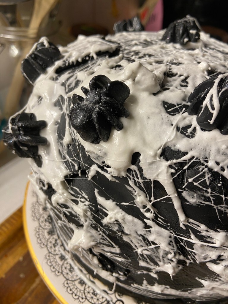 Detailaufnahme: Eine Schwarzer Fondant, Spinnweben aus Marshmallowfäden, schwarzsilberne Zuckerspinnen. Eine Spinne in Großaufnahme in der Mitte des Bildes.
