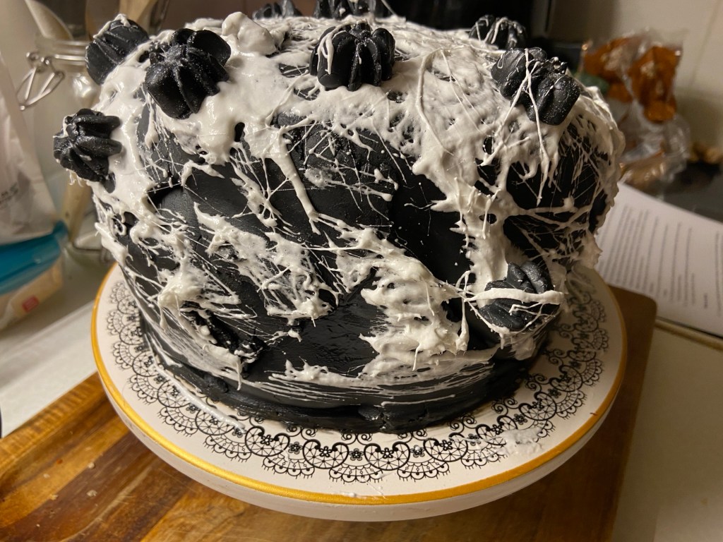 Eine Torte: Schwarzer Fondant, Spinnweben aus Marshmallowfäden, schwarzsilberne Zuckerspinnen. Alles sieht sehr selbstgemacht und ein wenig schief aus.