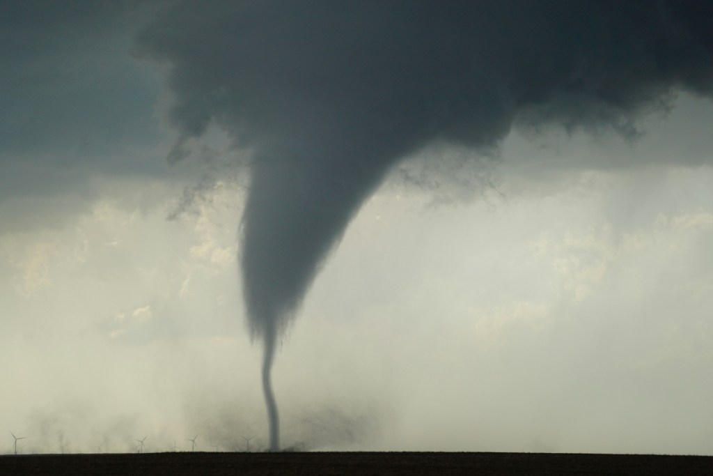 Bild in Grautönen: In der Ferne am Horizont stehen winzige Windräder. Darüber erhebt sich eine gigantische Windhose, die oben in dunkelgrauen Sturmwolken endet.