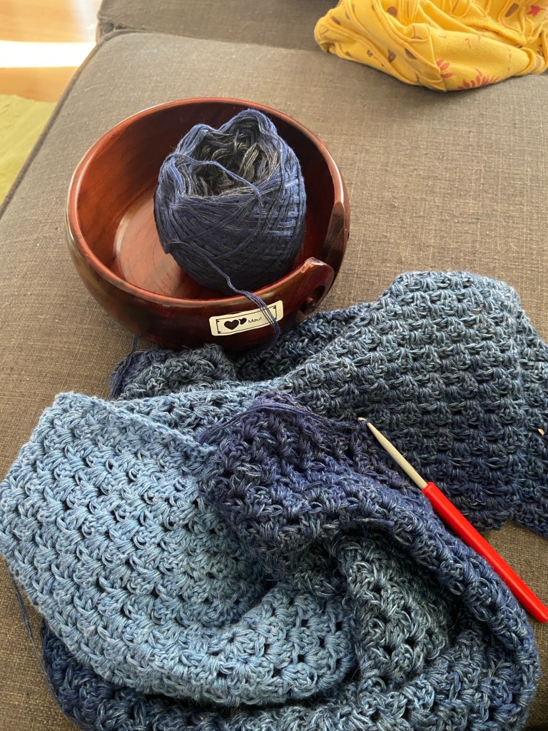 Häkelwerk: Dreieckstuch in verschiedenen Blautönen. Das unfertige Tuch liegt zusammengeknüllt auf dem grauen Sofa, das noch nicht verarbeitete Garn liegt in einer Holzschale.