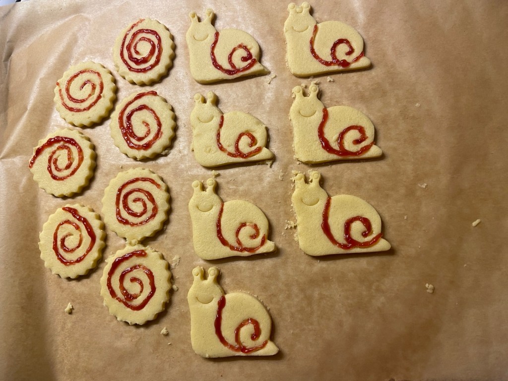 Ungebackene Plätzchen. Links rund ausgestochene Kekse mit gezacktem Rand, auf die rote Spiralen mit Gelee gemalt sind. Rechts fröhliche Schnecken, bei denen einige Details rot nachgezogen sind.