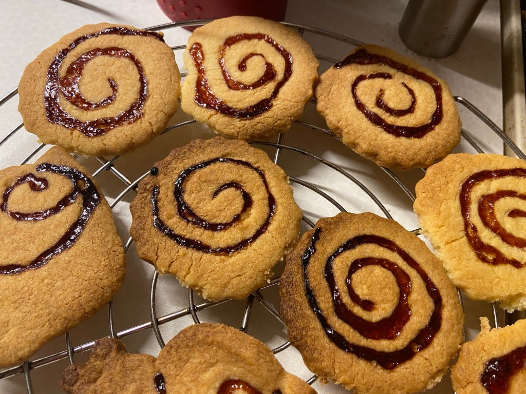 Gebackene Spiralkekse, leicht gebräunt.: Runde Kekse mit gezacktem Rand und eingesunkener roter Geleespirale 