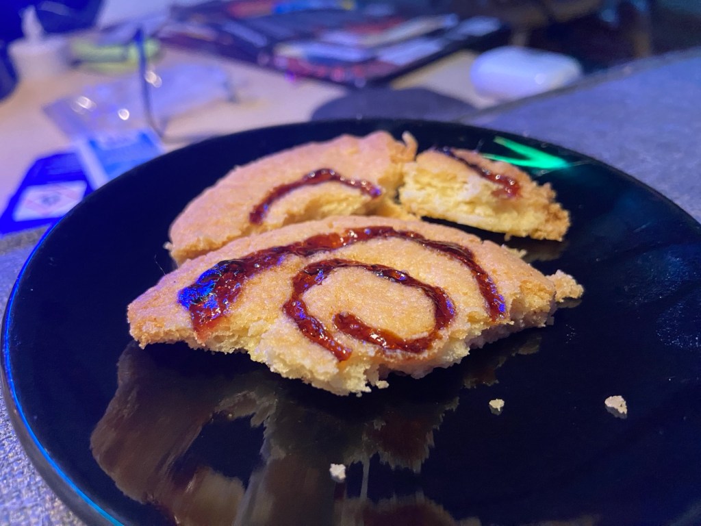 Ein zerbrochener Spiralkeks auf einem schwarzen Tellerchen. Man sieht im Querschnitt, dass die rote Spirale nicht bis zum Boden des Kekses reicht, sondern nur eingekerbt war.