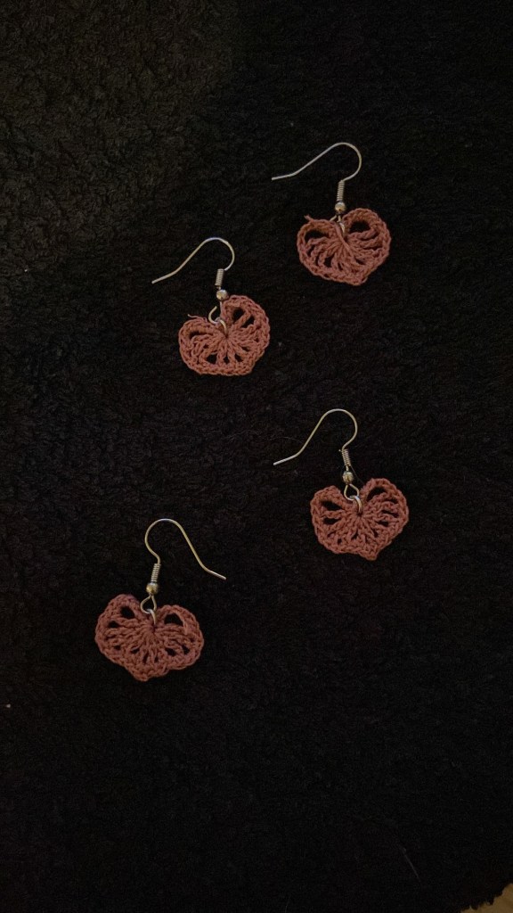 Schwarzer Hintergrund, darauf liegen vier Hänge-Ohrringe: Filigrane Herzen aus rosa Stickgarn.