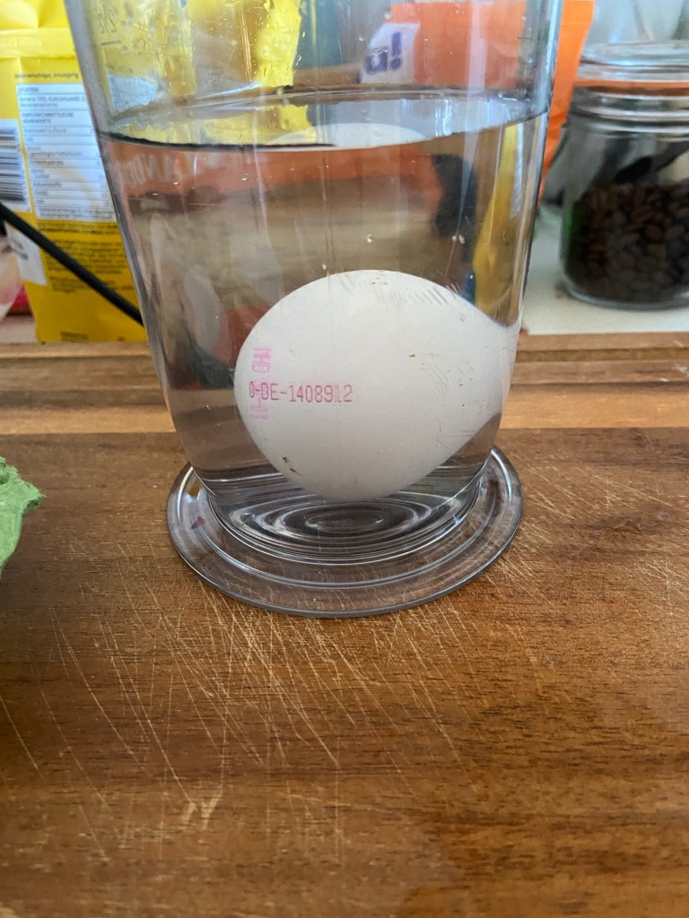 Messbecher mit Wasser auf hölzerner Anrichte. In dem Messbecher liegt ein weißes Hühnerei.