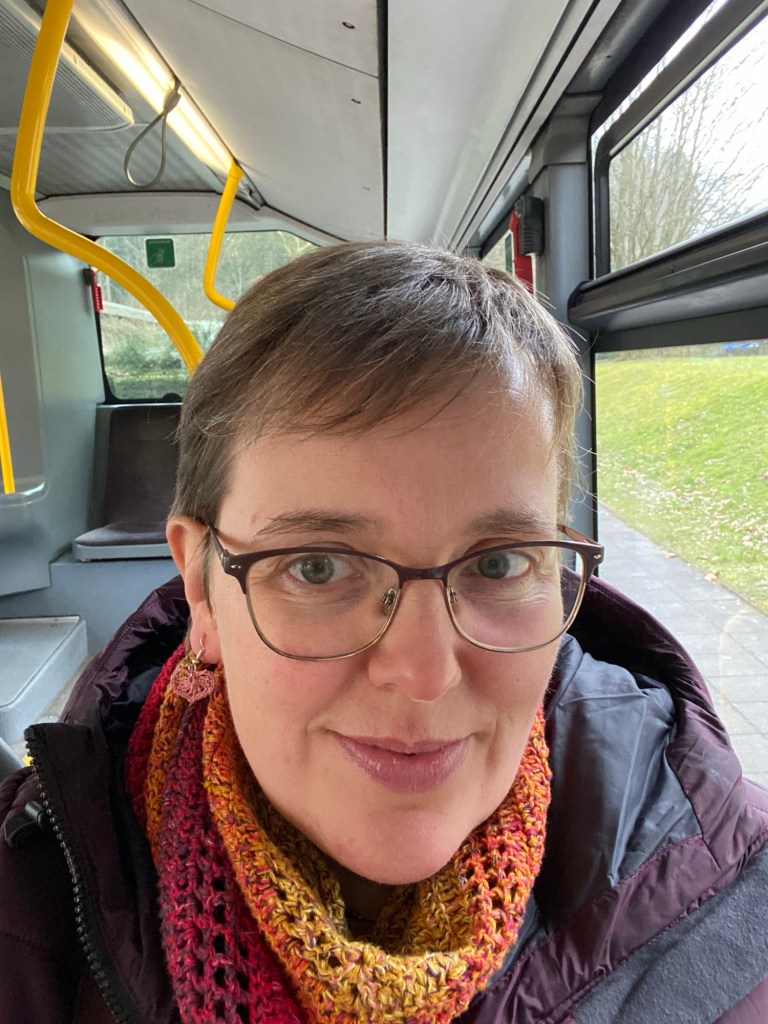 Selfie im Bus: Autorin mit kurzem blondem Haar, großer Brille und herbstfarbenem Häkelschal. An einem Ohr erkennt man einen winzigen gehäkelten Ohrring aus rosa Garn.
