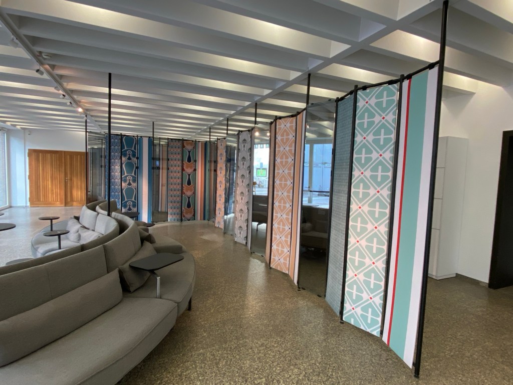 Im MAKK - schmale Panelen mit geometrischen Mustern oder Spiegeln bilden eine Ecke, darun steht ein geschwungenes graues Sofa im Raum.