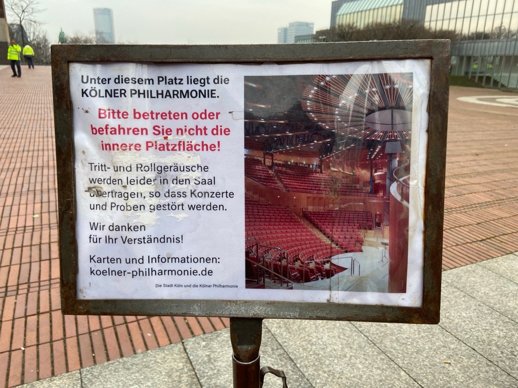 Dieses Schild steht vor dem Platz über der Kölner Philharmonie. Es sieht ein wenig mitgenommen aus. Zusammengefasst steht da, man solle den Platz bitte nicht betreten, weil man die Schritte im Saal unter dem Platz als störend höre.