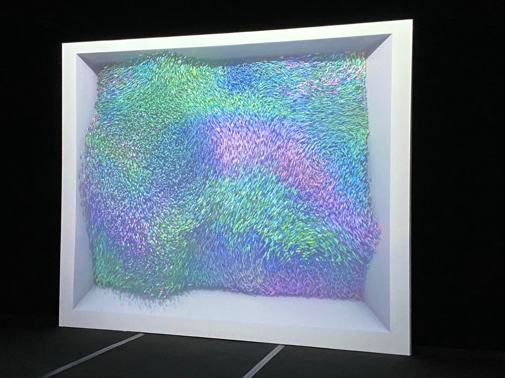 Schwarzer Raum, große weiße Leinwand. Auf der Leinwand sieht man viele winzige Pünktchen und Striche in Grün, Türkis, Blau und Violett, die sich zu bewegen scheinen wie ein Weizenfeld im Wind. Das Bild wirkt dreidimensional und plastisch.