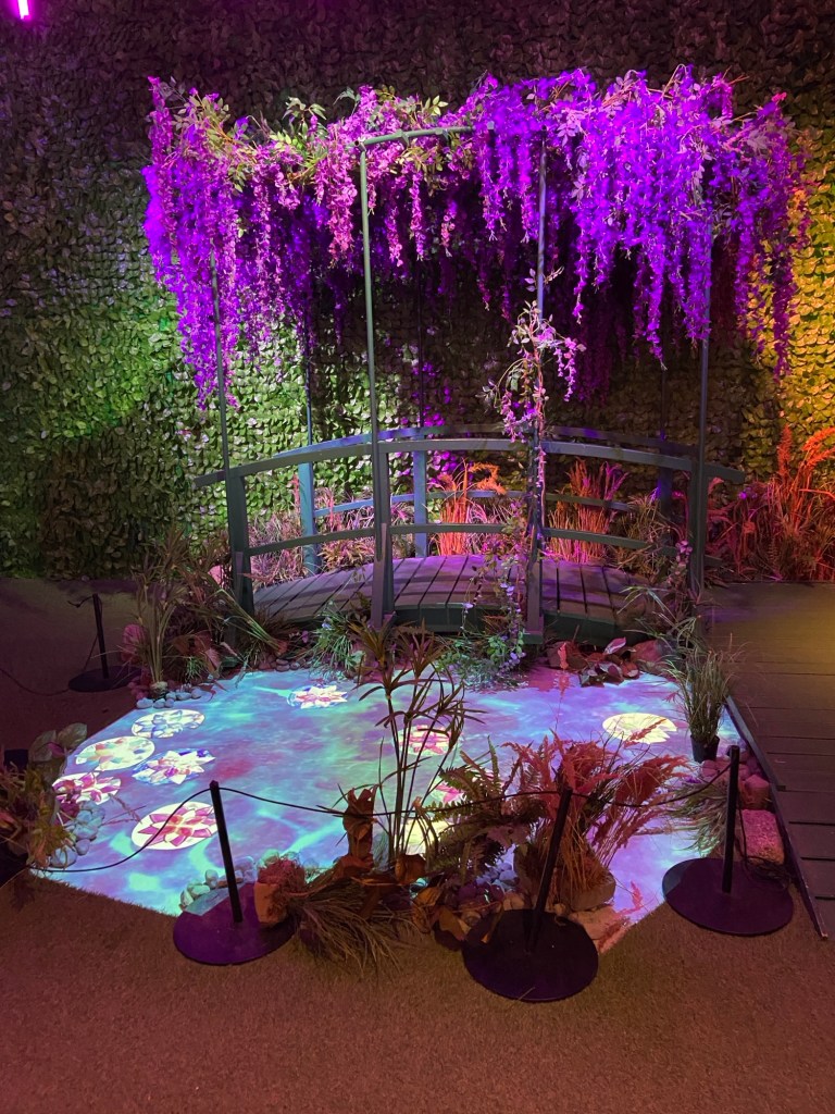 Eine mit Wisterien bewachsene Holzbrücke führt über einen projizierten Teich voller gemalter Seerosen. Der Hintergrund besteht aus einer mit Efeu bespannten Wand.