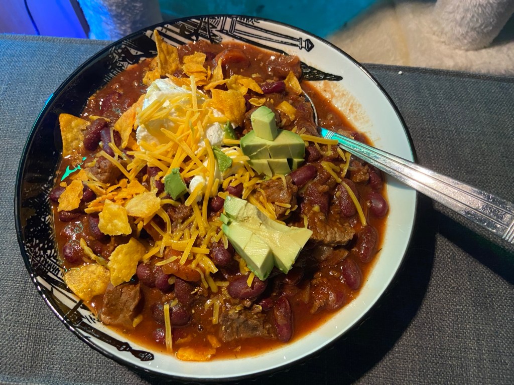 Ein Teller Chili con Carne mit Avocado-Stücken, Speisequark, geriebenem Käse und Nacho-Chips.