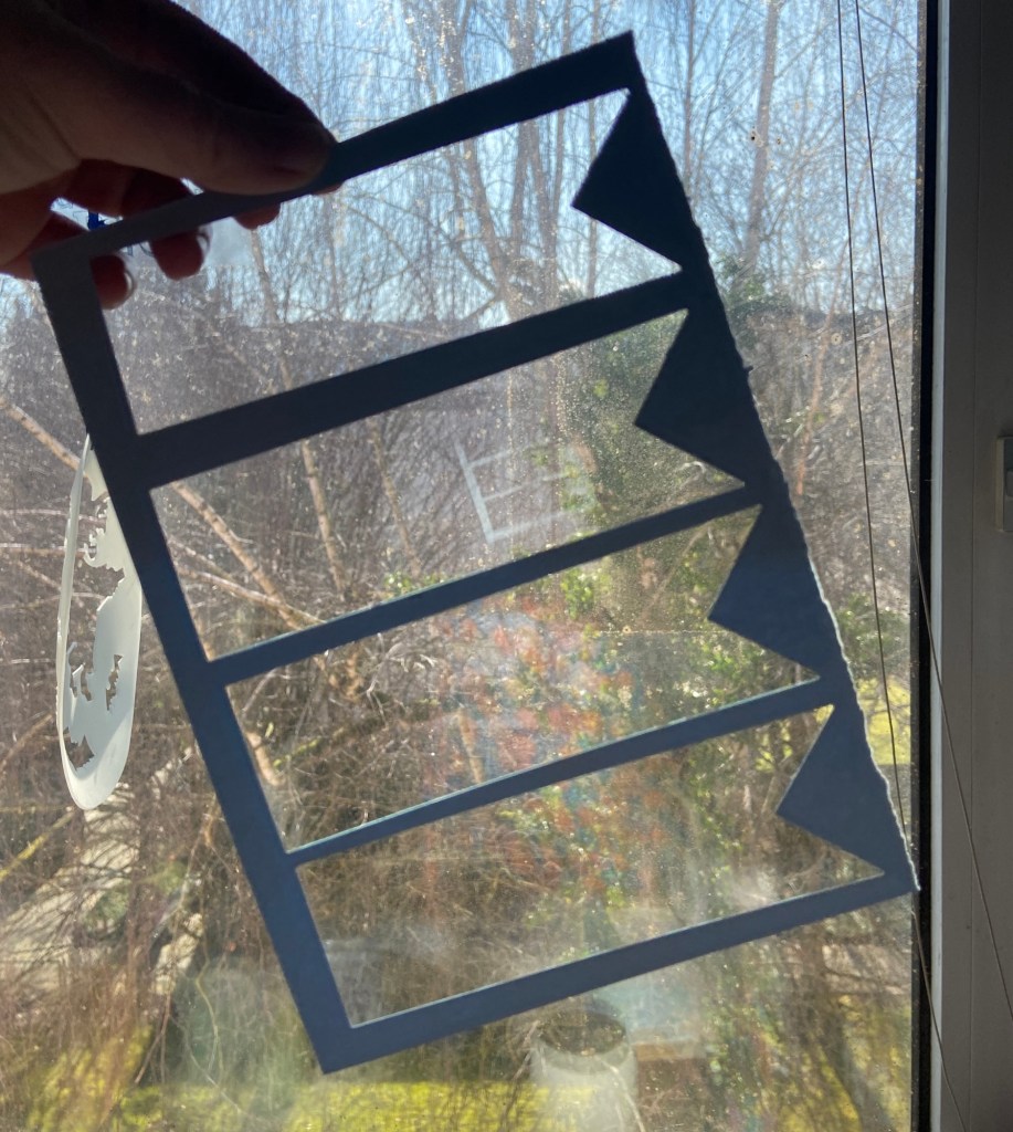 Vor einem Fenster hält eine Hand ein Stück blauen Tonkarton hoch, aus dem vier Lesezeichen ausgestanzt wurden.