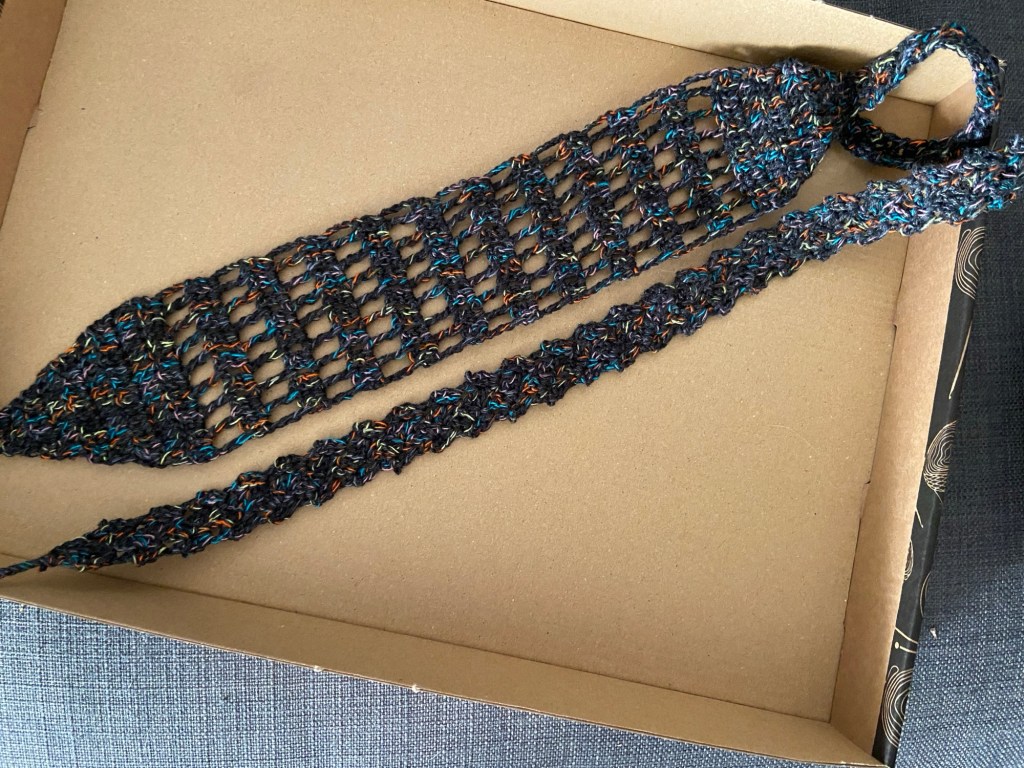 In einem Pappdeckel liegen ein breites und ein schmales Stirnband aus schwarz-buntem Garn, beide gehäkelt.