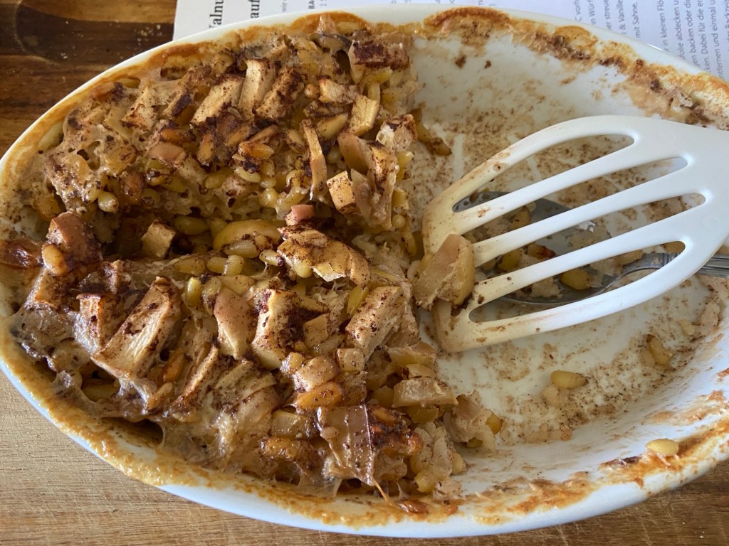 Eine ovale Auflaufform, noch halb gefüllt mit Milchreisauflauf mit Äpfeln und Pinienkernen. Die rechte Hälfte ist schon verputzt, da liegt ein weißer Pfannenwender.