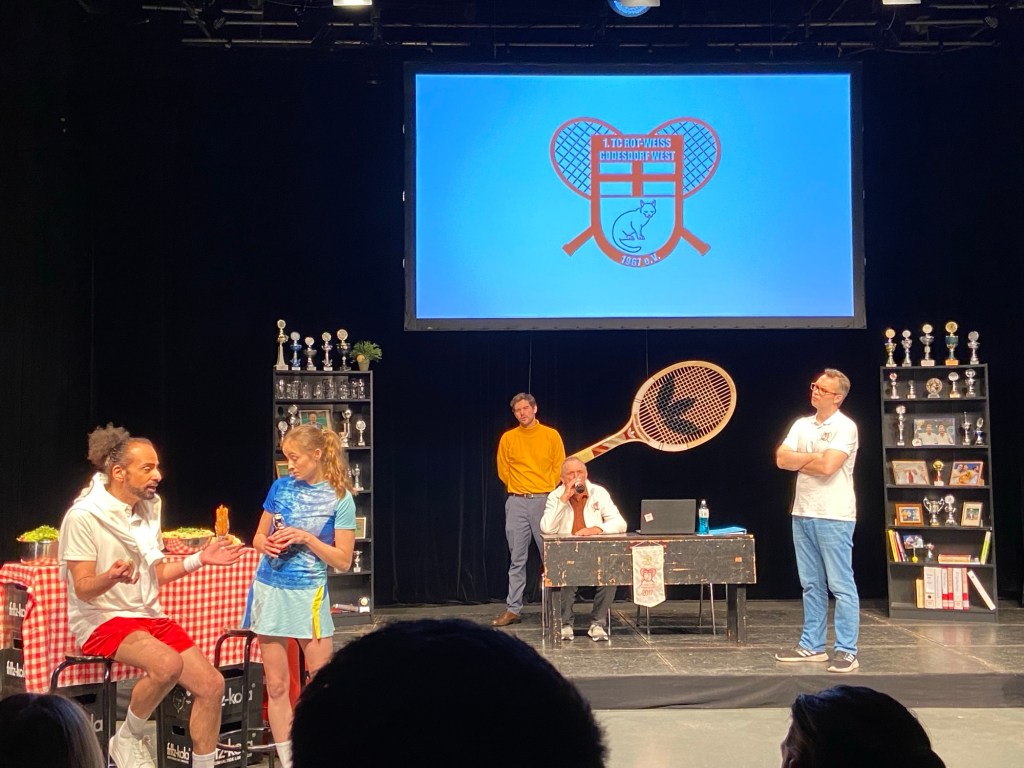Theater: Eine nachgestellte Mitgliederversammlung in einem Tennisverein. Vorn links sitzen ein Mann und eine Frau in Sportkleidung und unterhalten sich. Mittig-rechts im Bild ein Tisch, an dem ein älterer Herr neben einem Notebook sitzt. Hinter ihm steht ein Mann in einem gelben Rollkragenpullover. Rechts davon, weiter vorn im Bild, steht ein Mann mit weißen T-Shirt und verschränkten Armen. Links und rechts vom Tisch stehen Regale mit Trophäen, hinter dem Tisch ist das Logo des Vereins auf eine weiße Fläche projiziert: Gekreuzte Tennisschläger vor einem Wappen mit einem Kreuz und einer Katze. Das Wappen ist rot auf weißem Grund.