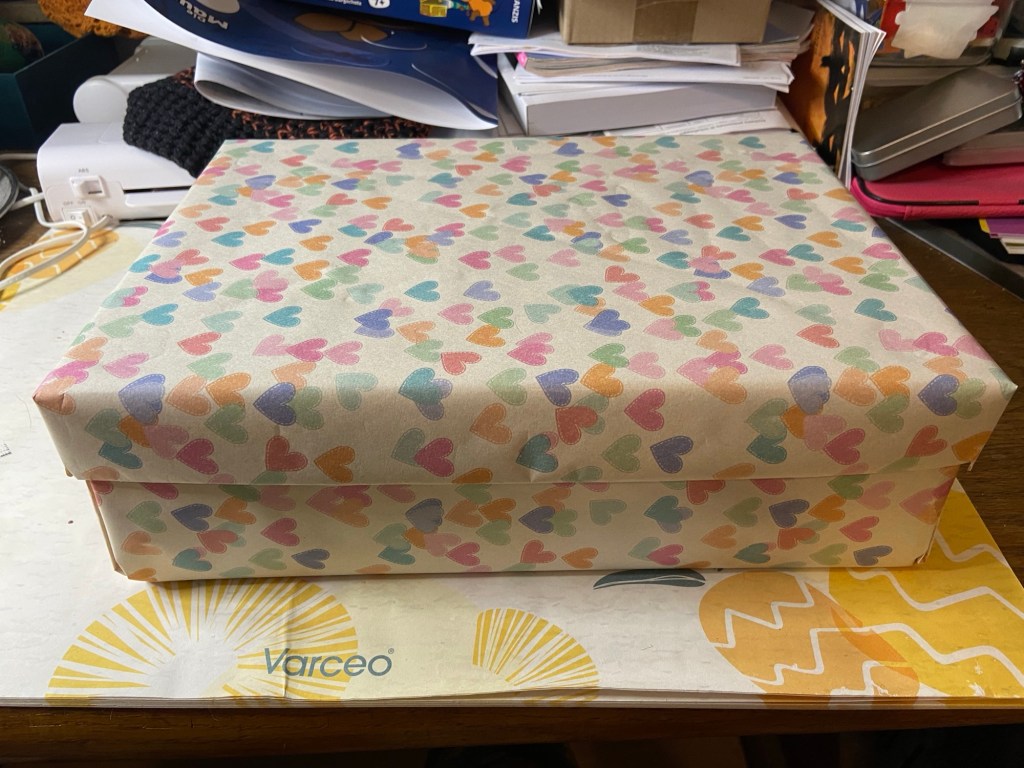 Eine in Geschenkpapier mit bunten Herzchen einigermaßen ordentlich eingekleidete Pappbox auf einem chaotischen Schreibtisch. 
