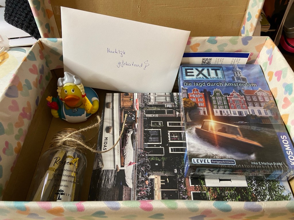 Der Inhalt der Box. Von links nach rechts - eine "Frau Antje"-Gummiente, ein Umschlag mit der Aufschrift "Hartelijk gefeliciteerd", ein Amsterdam-Bildband und ein Amsterdam-Exit-Room-Spiel. Vorne links eine Glasflasche mit der Reisekasse. Ganz hinten erkennt man zwei Tickets für ein Event (die haben mit dem eigentlichen Geschenk nichts zu tun).