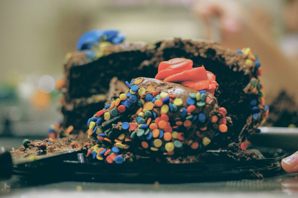 Im Hintergrund eine angeschnittene Schokoladentorte mit vielen bunten Zuckerstreuseln und bunten Buttercreme-Rosetten. Im Vordergrund ein einzelnes Stück Torte, leicht verunfallt und zermatscht.