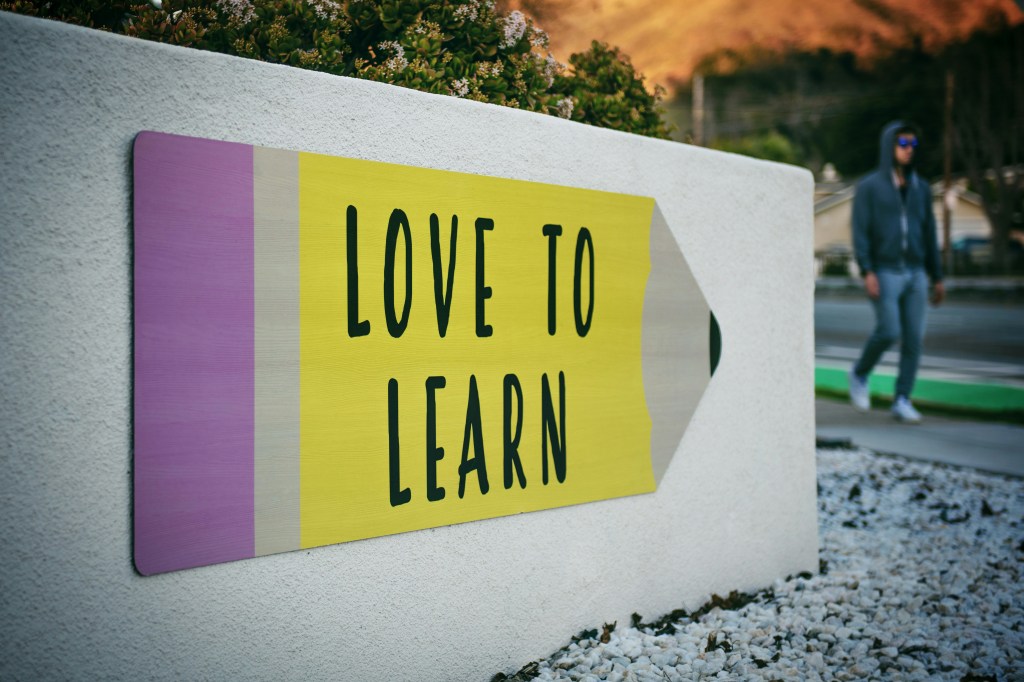 An einer etwa hüfthohen, weiß getünchten Mauer hängt ein großes Schild in Form eines Bleistifts mit der Aufschrift: Love to learn. 