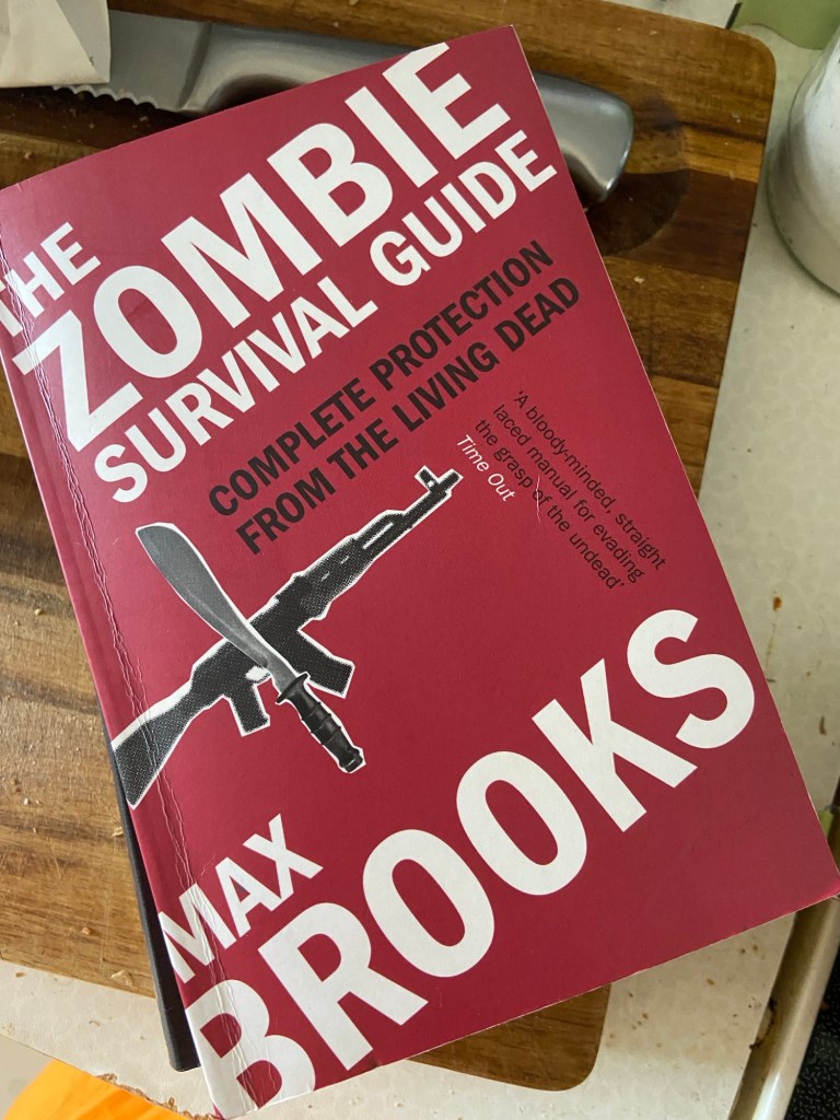 Buchcover: "The Zombie Survival Guide - Complete Protection from the Living Dead - A bloody-minded, straight laced manual for evading the grasp of the undead (Time Out) - Max Brooks". Das Cover ist rot mit weißer und schwarzer Schrift und collagen-artiger Anordnung von einem Maschinengewehr und einer Machete (in Schwarz-Weiß und körniger Auflösung). Das Buch liegt auf einem Küchenbrett, oben am Rand erkennt man noch ein Brotmesser.