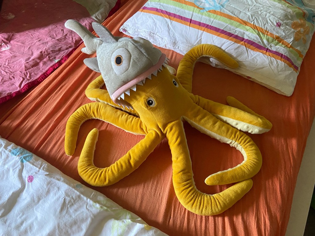 Auf einem bunten Bett sitzt ein orangefarbener Plüsch-Oktopus, der einen Hai als Hut trägt. 
